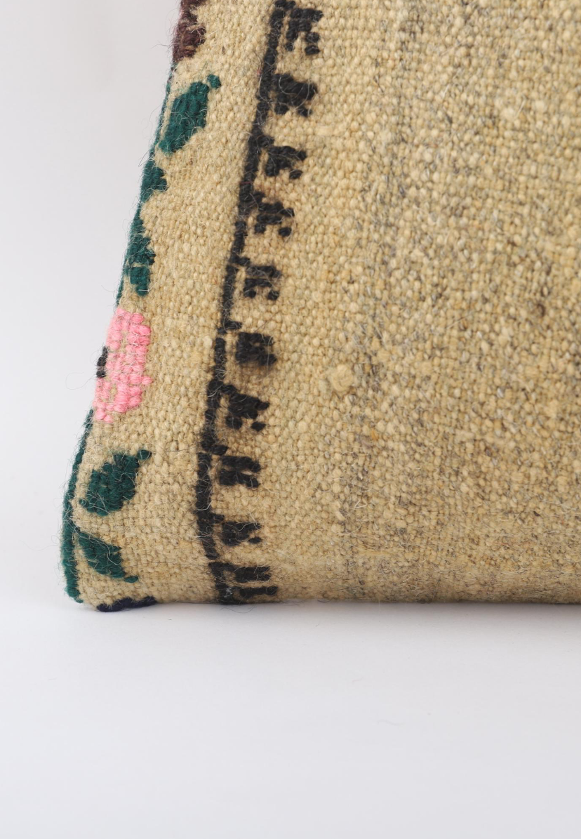 A close-up of a woven textile or rug corner showing a beige base with a black geometric border, green and pink floral accents, and a textured natural fiber surface.