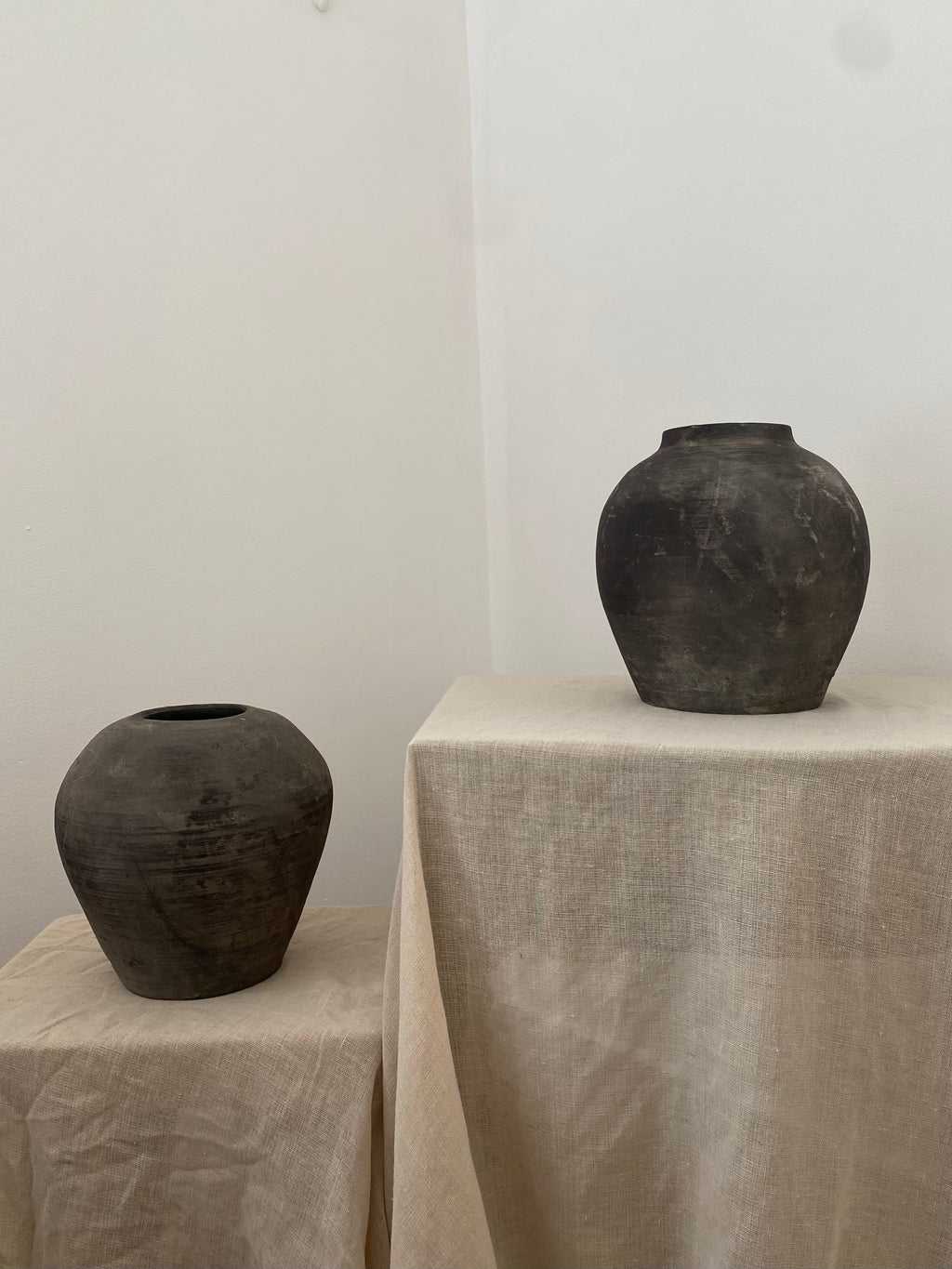 Two black antique terracotta clay pots on beige linen against a white wall