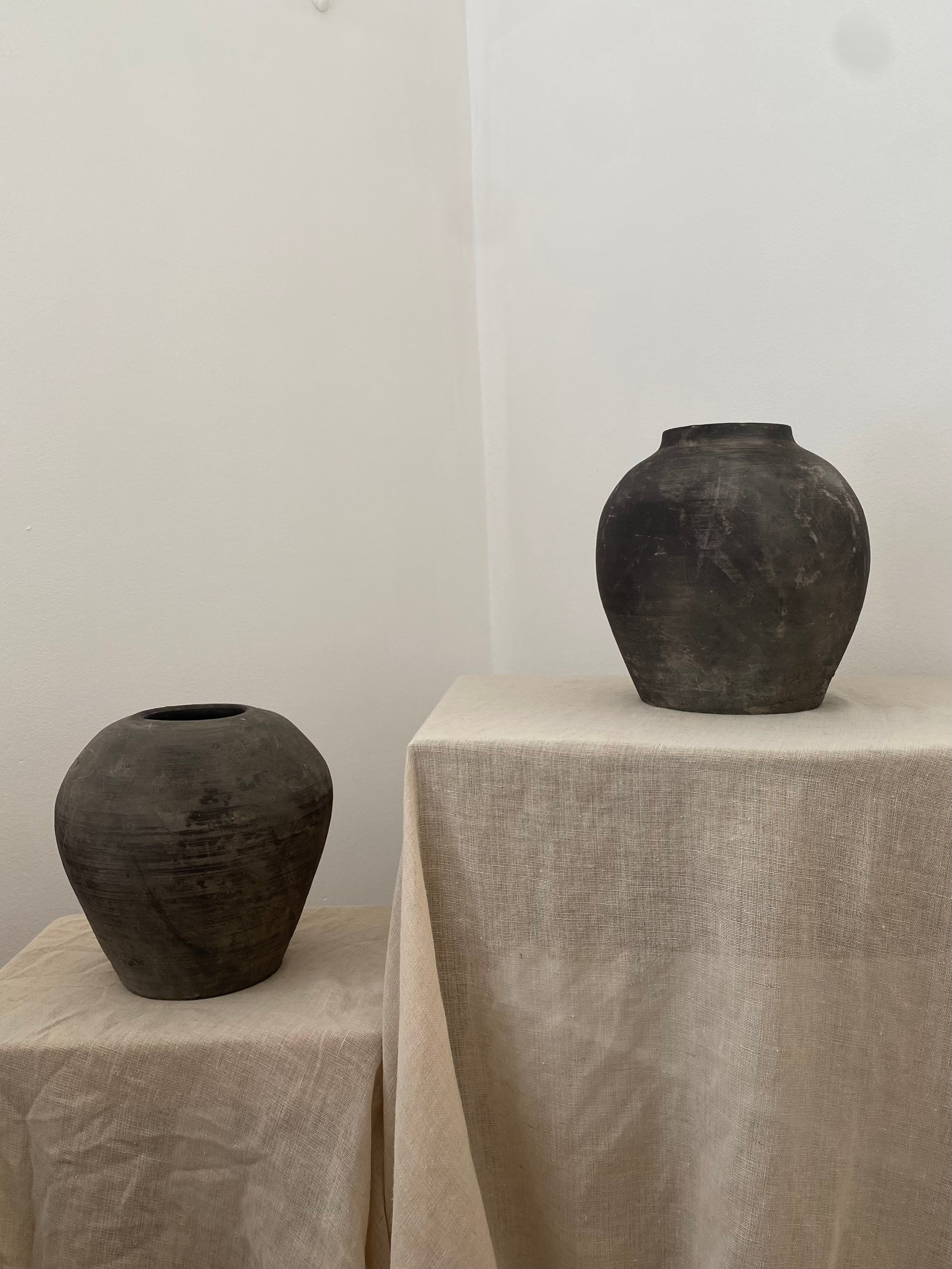 Two black antique terracotta clay pots on beige linen against a white wall