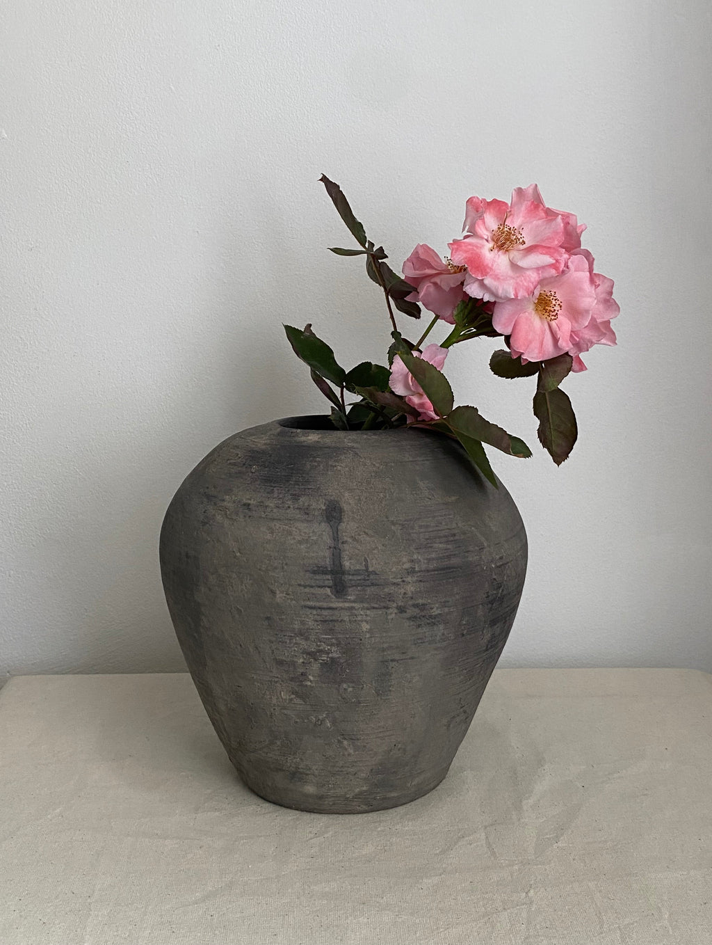 A dark grey antique clay pot with pink roses and green leaves, set on a light beige fabric-covered surface against a plain light gray wall.