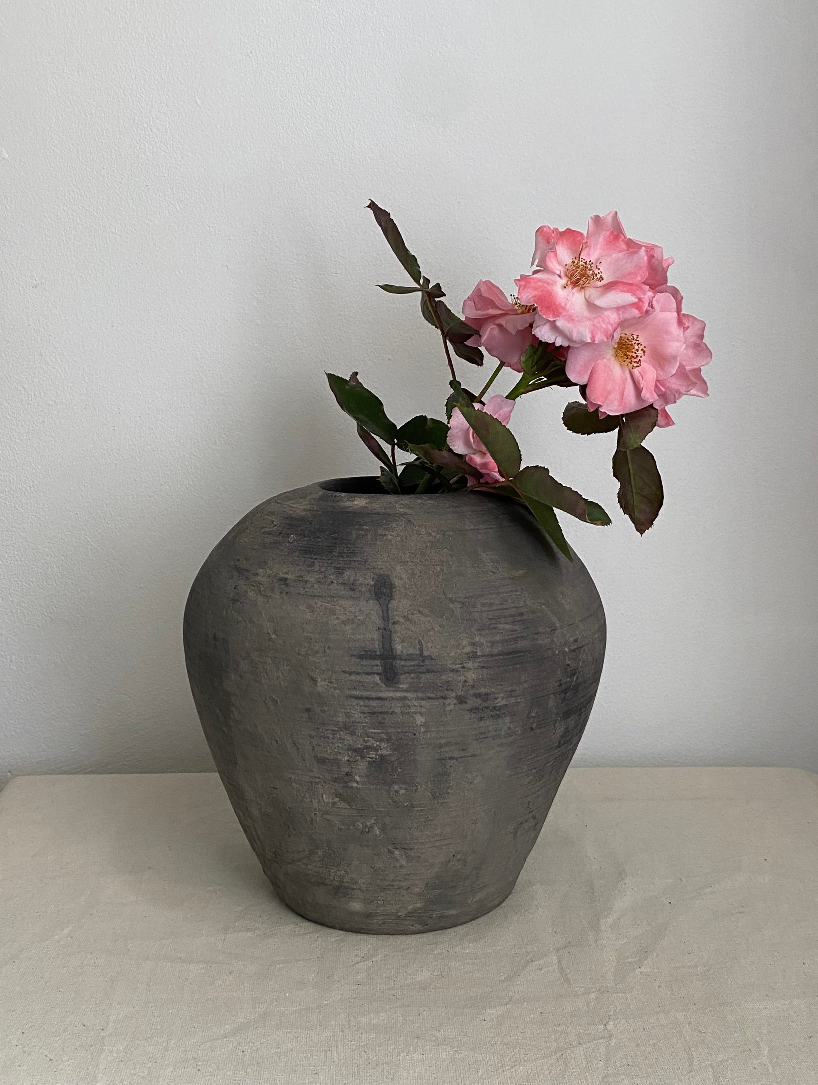 A dark grey antique clay pot with pink roses and green leaves, set on a light beige fabric-covered surface against a plain light gray wall.