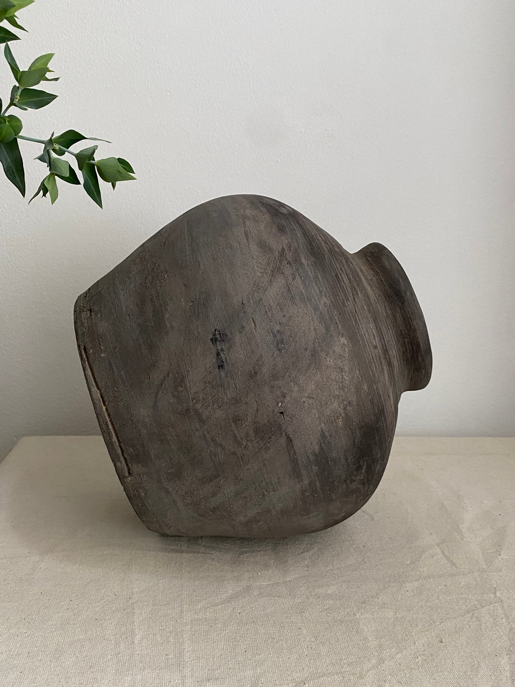 A large, grey rustic antique clay vessel with a wide rounded body and a short neck, resting on a light beige fabric-covered surface against a plain white wall, with a small green plant branch entering from the top left.