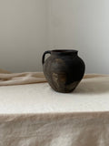 Black and brown antique clay pot leaning against a light-colored curtain on a neutral background