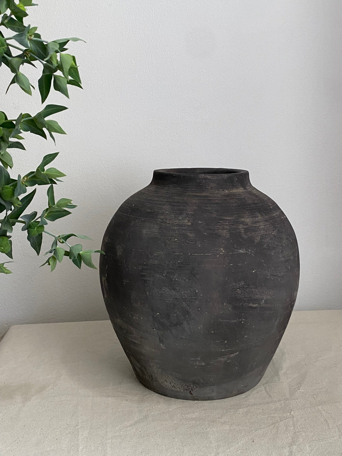 A large, dark charcoal antique terracotta clay pot with a textured matte finish, displayed on a light beige fabric-covered surface against a plain white wall, with green leaves from a plant entering from the left side.