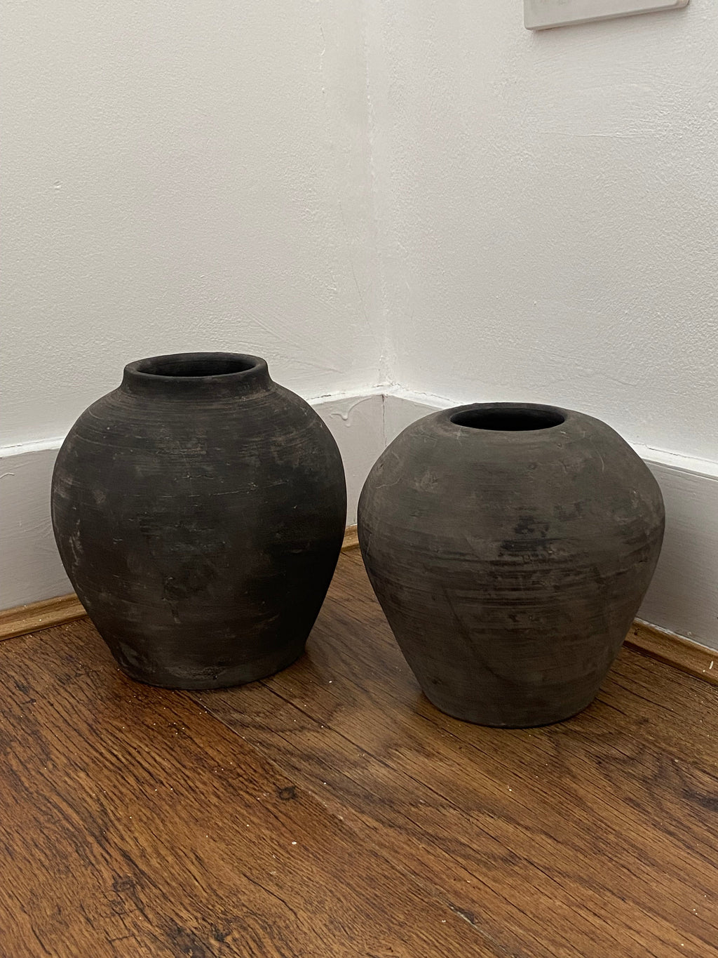 Two black terracotta clay pots on a wooden surface with a white wall background