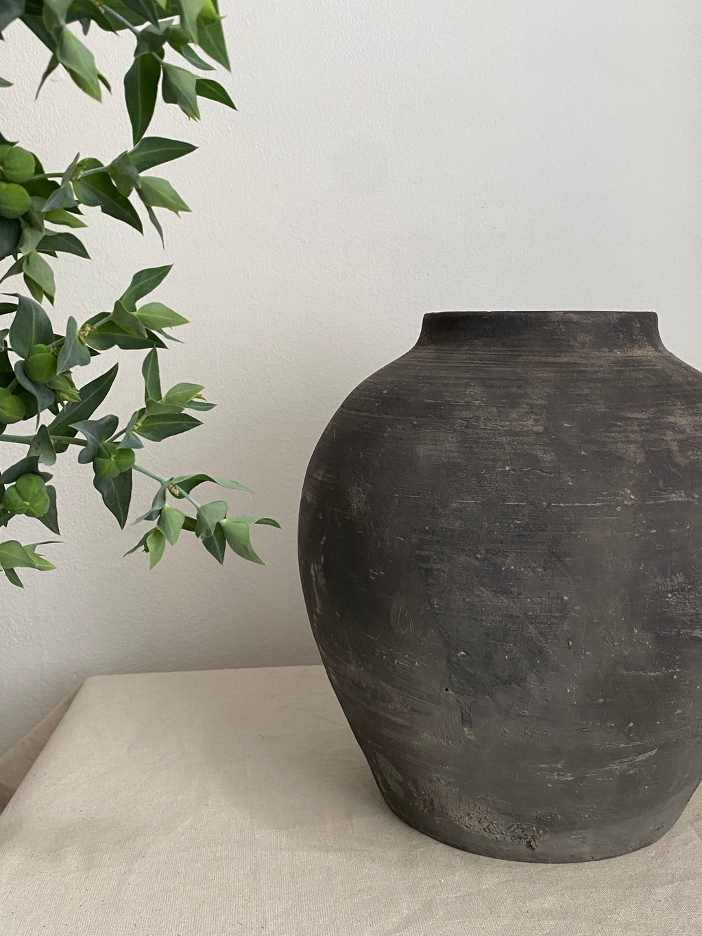 A large, dark charcoal terracotta clay vessel with a textured, rustic finish, sitting on a light beige fabric-covered surface against a plain off-white wall, with green plant leaves entering from the left side.