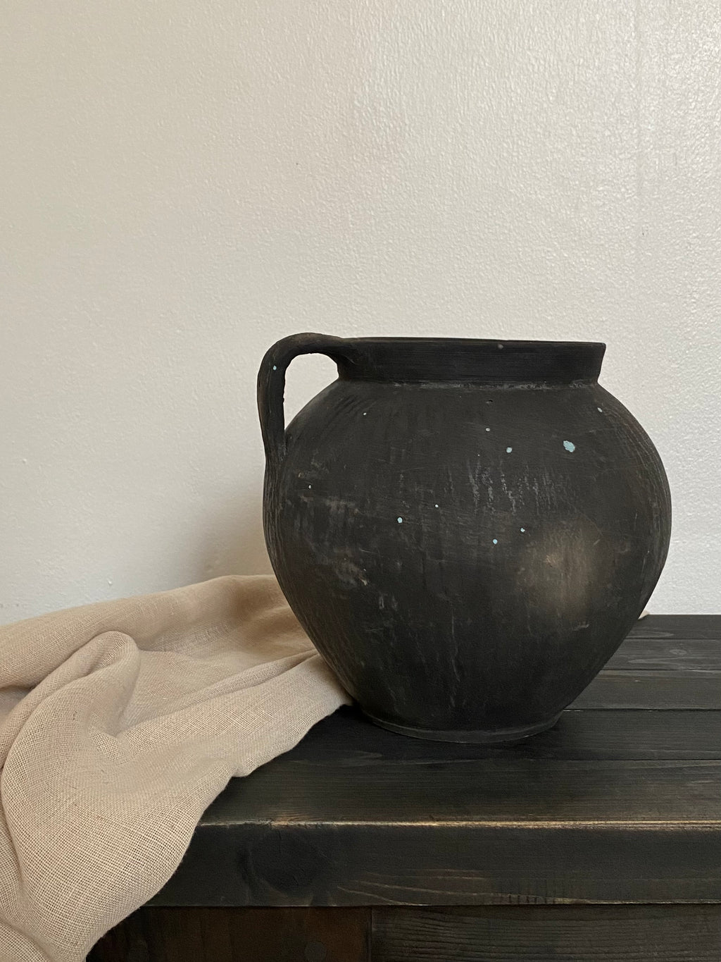 Black antique clay pot with a handle on a wooden bench against a white wall