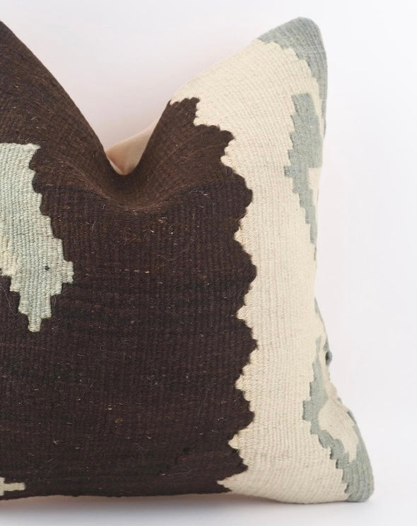 A close-up of a Anatolian Kilim woven throw pillow cover featuring a bold geometric pattern in dark brown, cream, and muted sage green on a white background.
