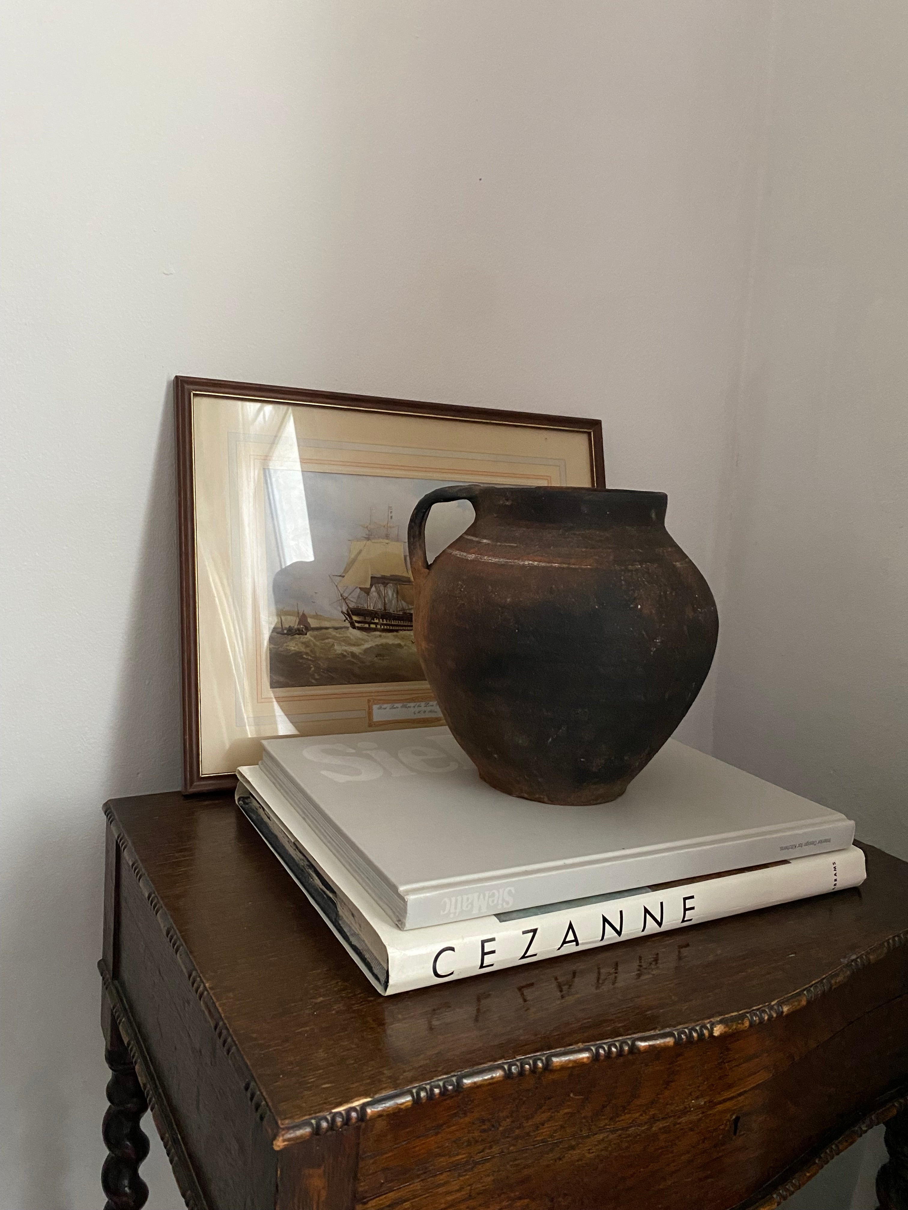 Antique terracotta clay pot on a wooden console with a stack of books and a framed picture in the background
