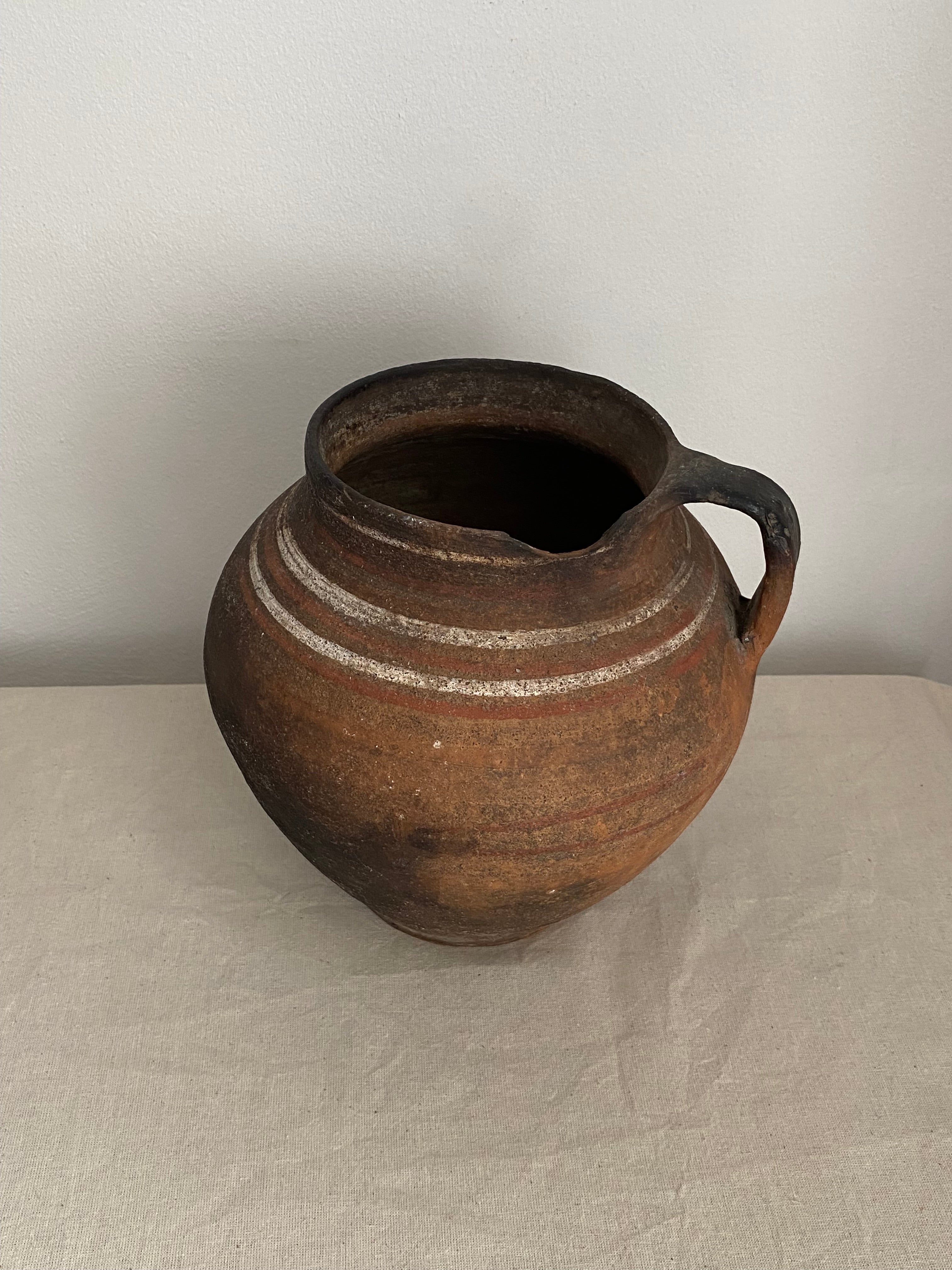 A rustic brown terracotta clay pot with a single loop handle, featuring two horizontal white painted stripes, shown against a neutral off-white background.