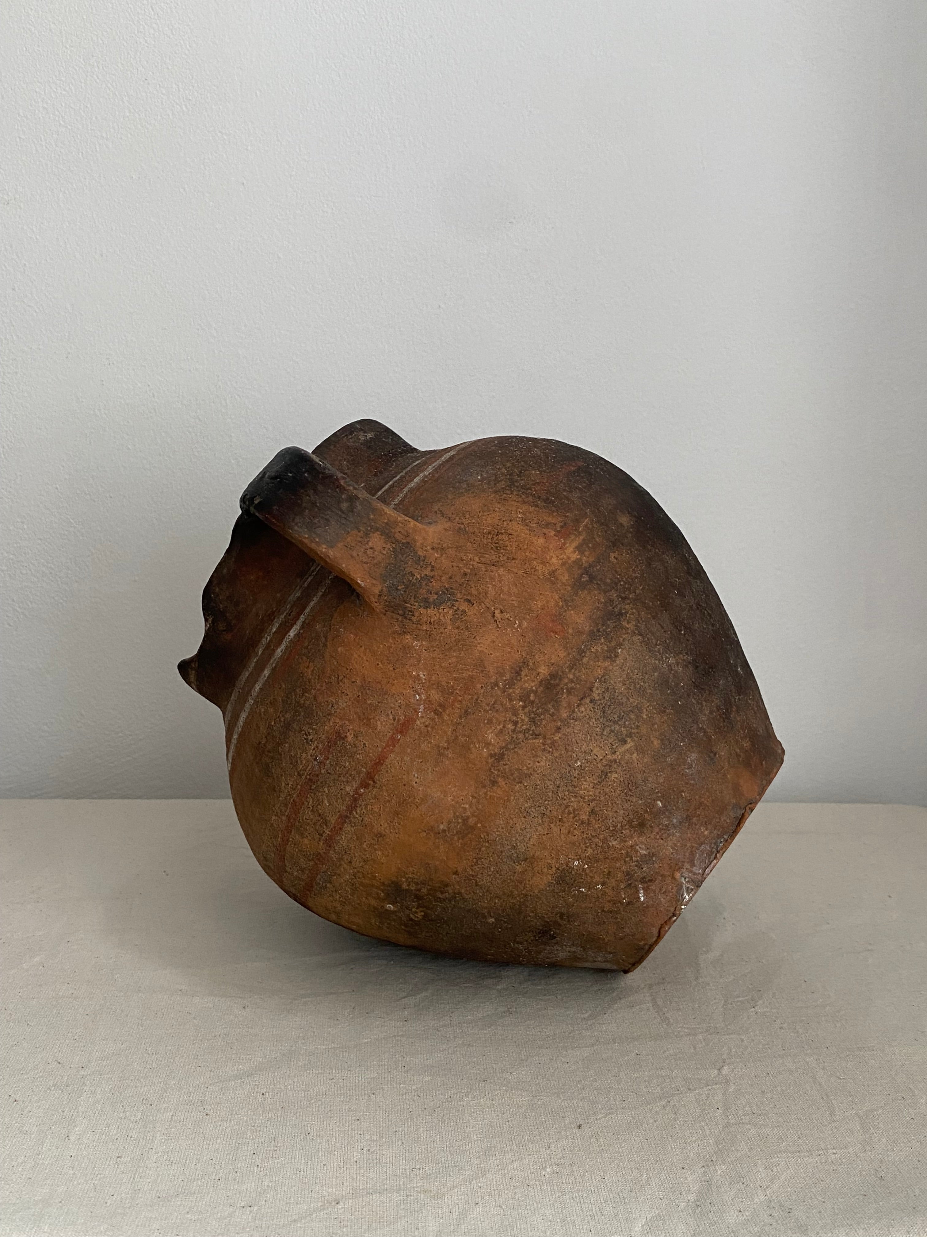 A rustic terracotta clay pot with a single handle, photographed against a neutral light gray textured background