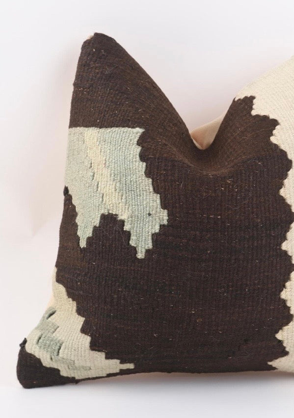 A close-up of a brown Turkish Kilim pillow cover  featuring a dark brown and cream geometric pattern with light sage green accents, set against a plain white background.