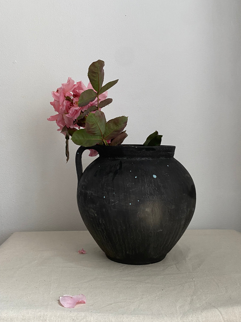 Black antique clay pot with a small side handle, holding pink flowers with green leaves, set against a plain off-white wall.