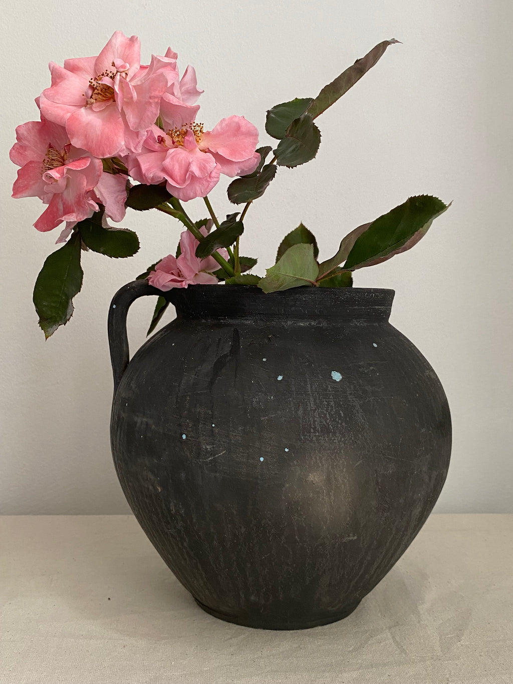 A rustic black terracotta clay pot with a small handle, filled with pink roses and green leaves, set against a plain off-white background.