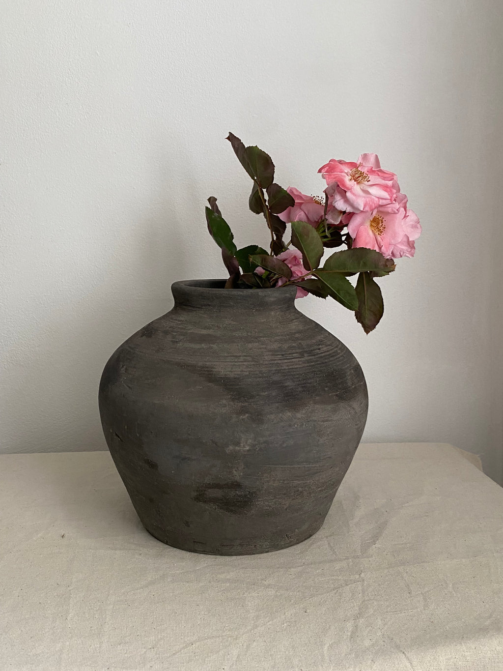 A dark grey rustic terracotta clay pot with a rounded bulbous shape, holding a pink flowering branch with green leaves, set on a light beige fabric-covered surface against a plain off-white wall.