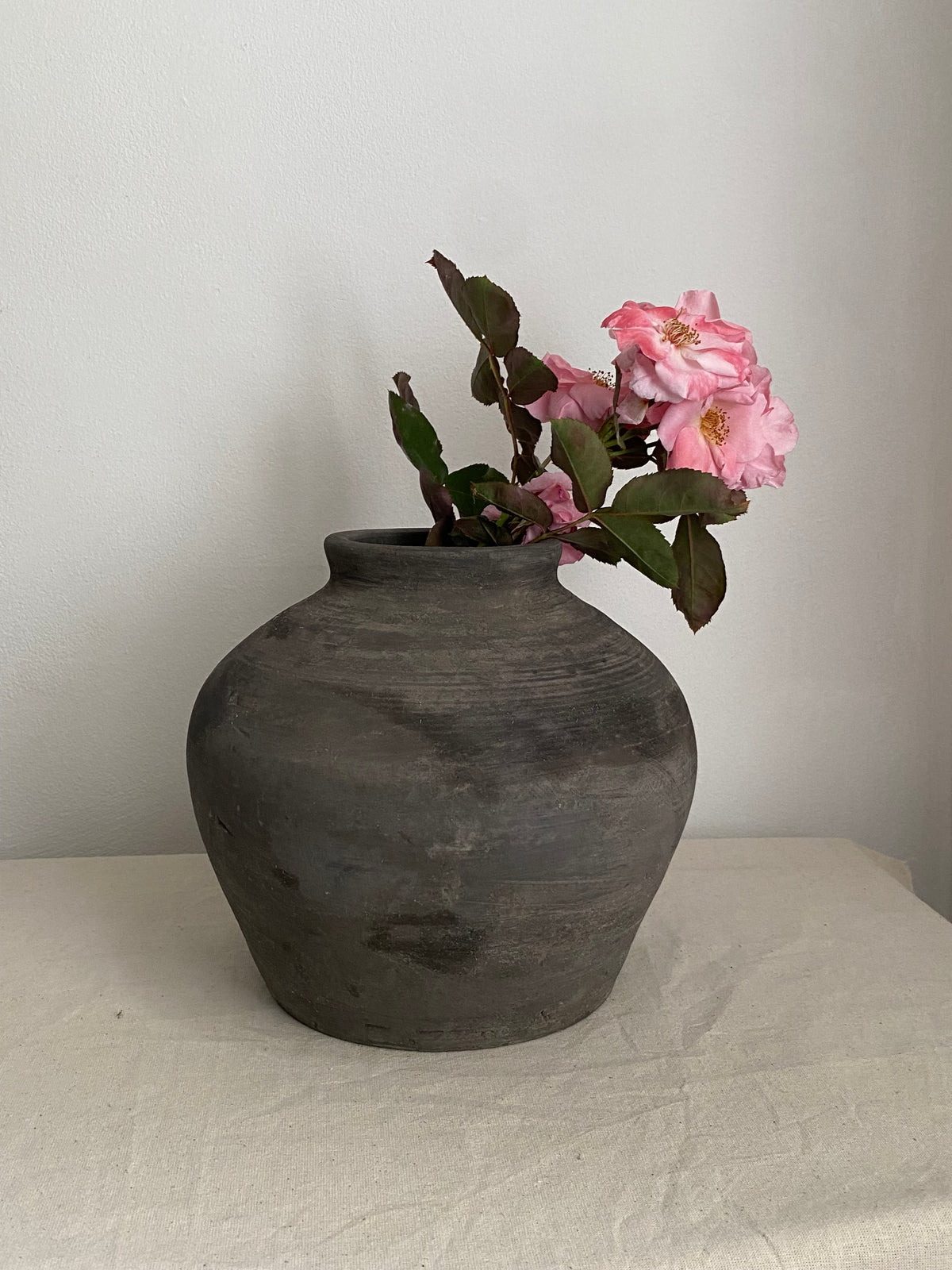 A dark grey rustic terracotta clay pot with a rounded bulbous shape, holding a pink flowering branch with green leaves, set on a light beige fabric-covered surface against a plain off-white wall.