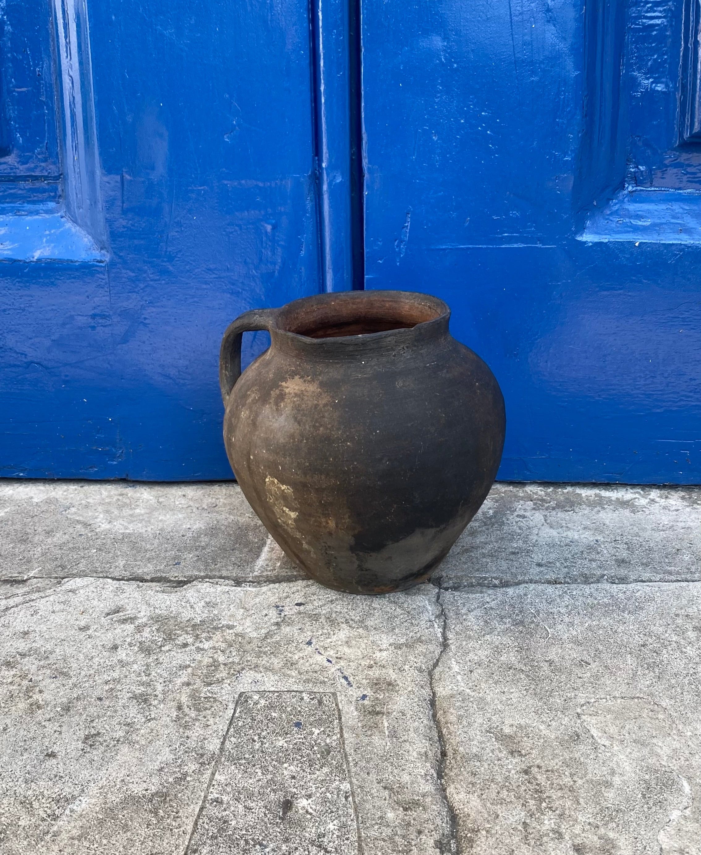 Black and brown rustic antique terracotta clay pot on a concrete surface with a blue metal door in the background