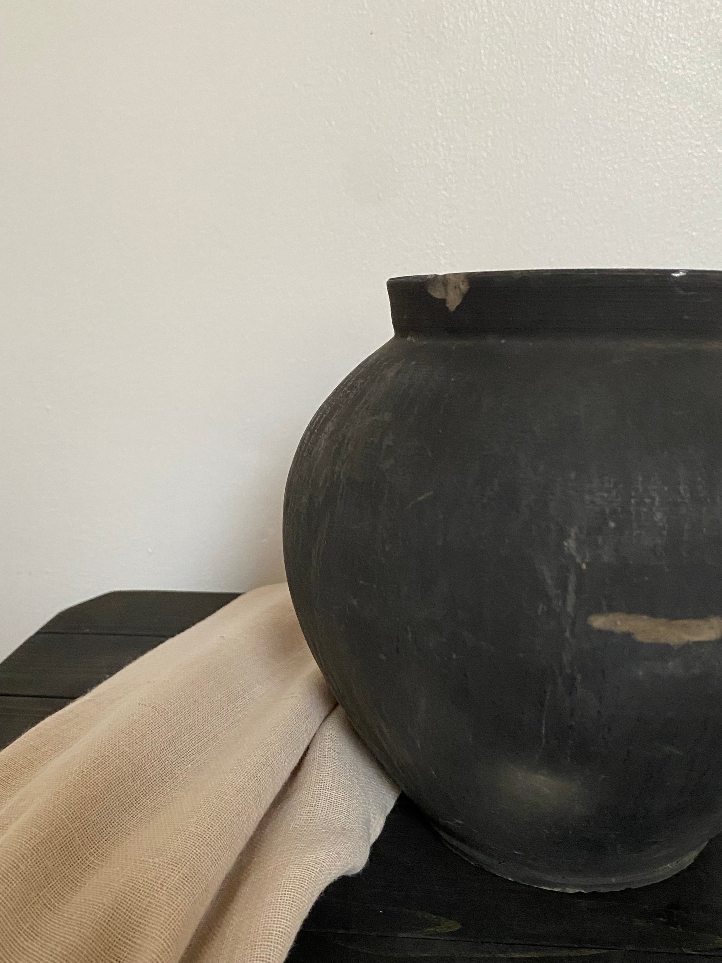 Close-up of a black antique clay pot on a wooden bench with draped beige fabric over it.