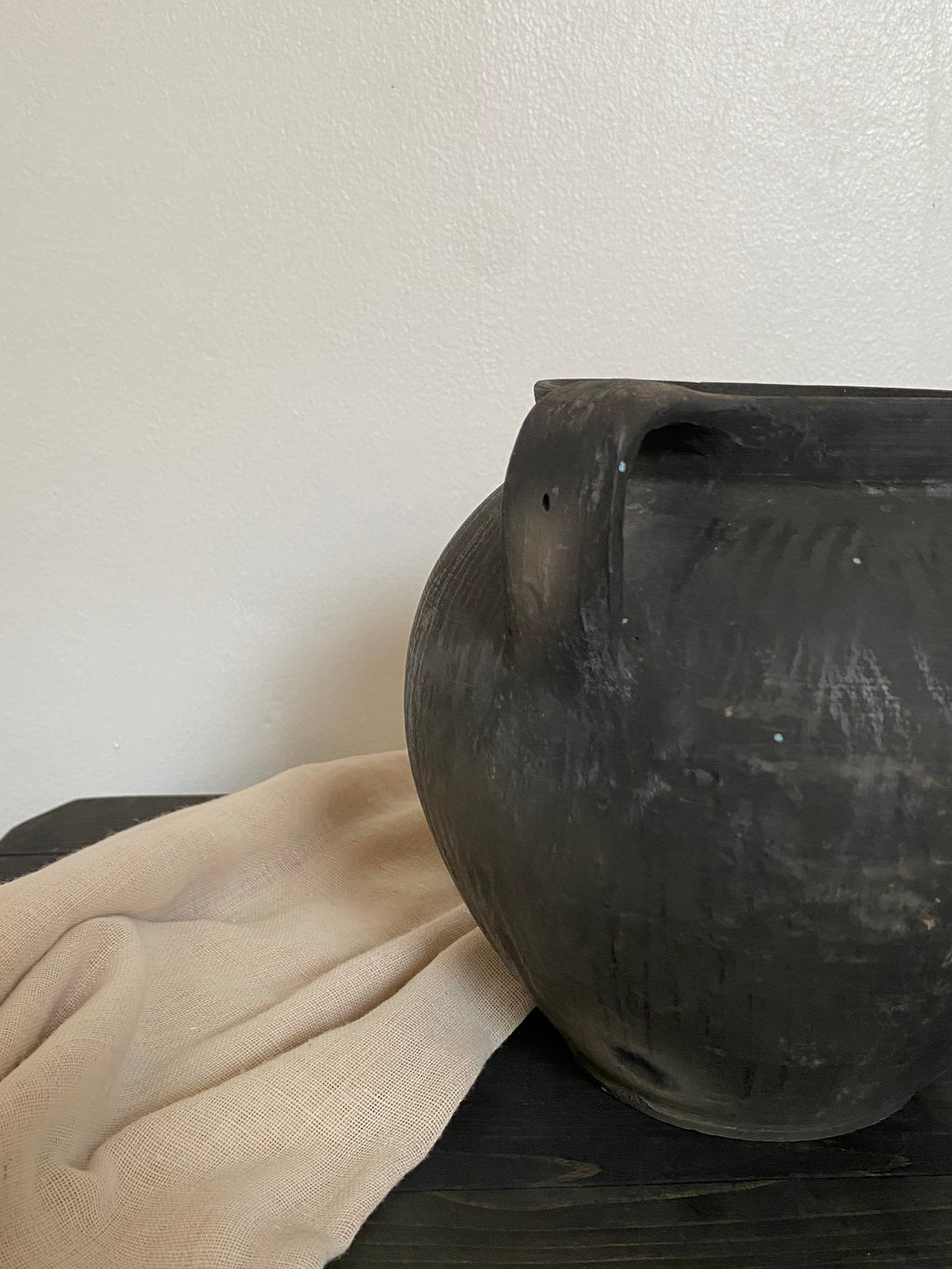 Close-up of a black antique clay pot  with a beige cloth on a wooden bench and a light neutral background 