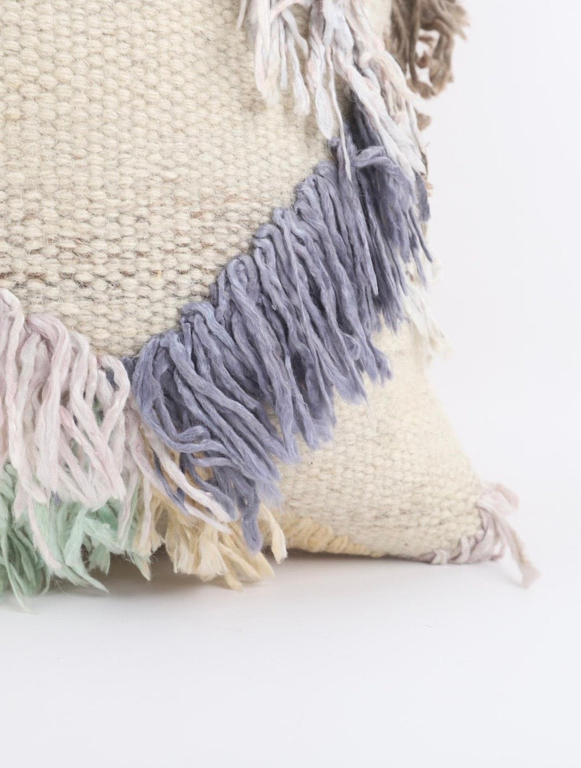 A close-up of a Turkish Kilim pillow cover throw pillow in a warm cream colour with layered fringe tassels in dusty blue, pale pink, and mint green, set against a clean white background.