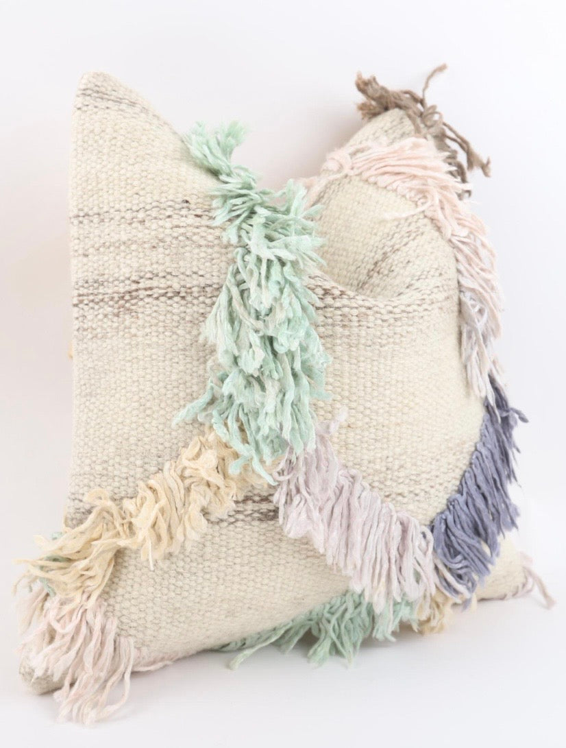 A square woven Anatolian Turkish Kilim pillow cover in a natural beige burlap-like fabric, decorated with soft pastel fringe tassels in mint green, lavender, cream, and dusty blue, set against a plain white background.