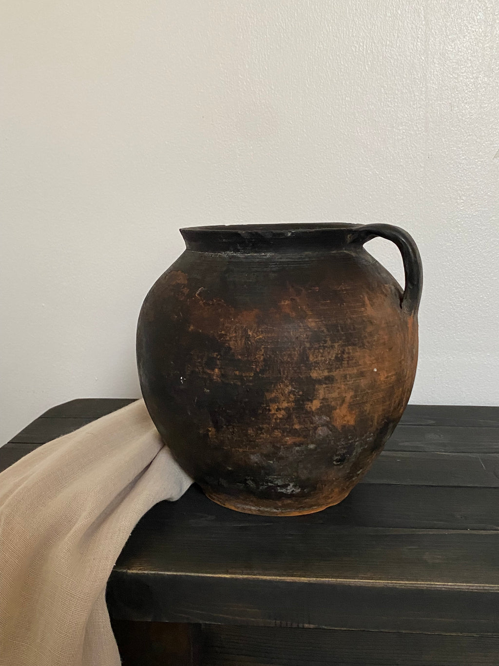 Rust-coloured antique clay pot with a handle against a white wall.