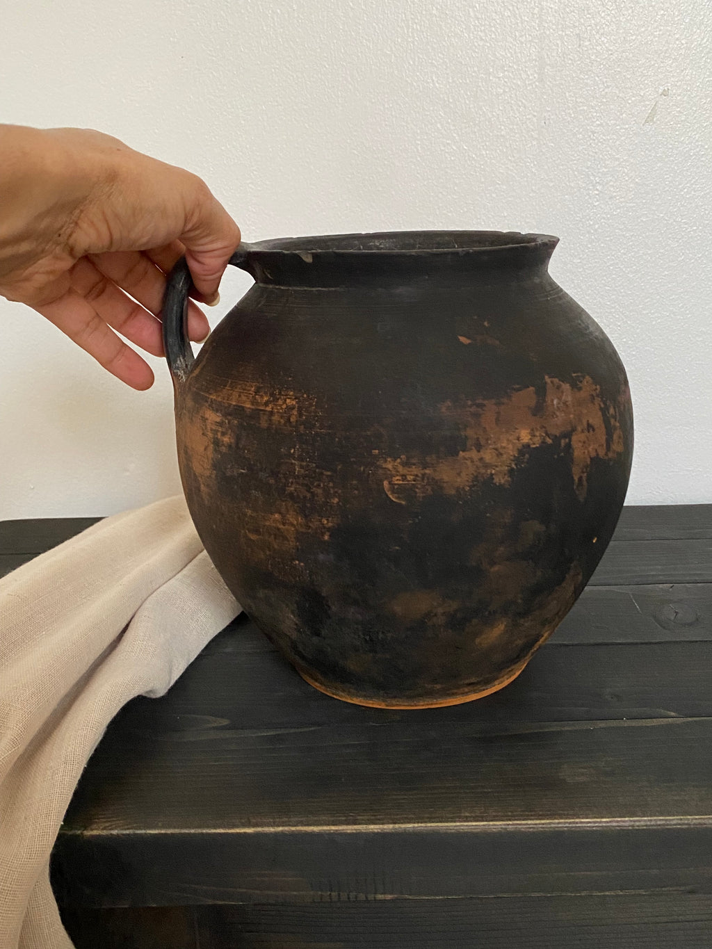 Hand holding an antique terracotta clay pot against a white wall and dark wooden surface.