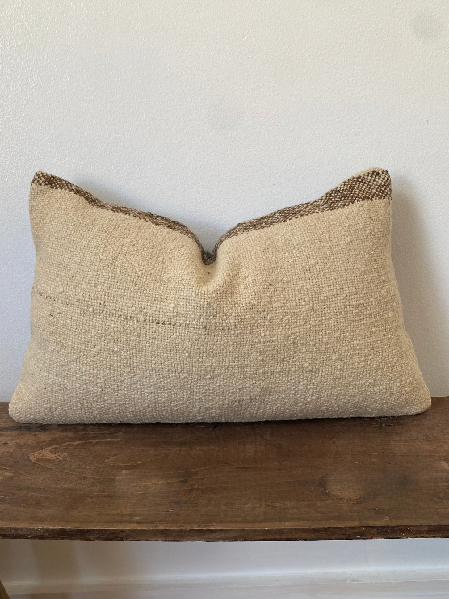 Textured beige pillow with brown trim on a wooden surface against a white wall