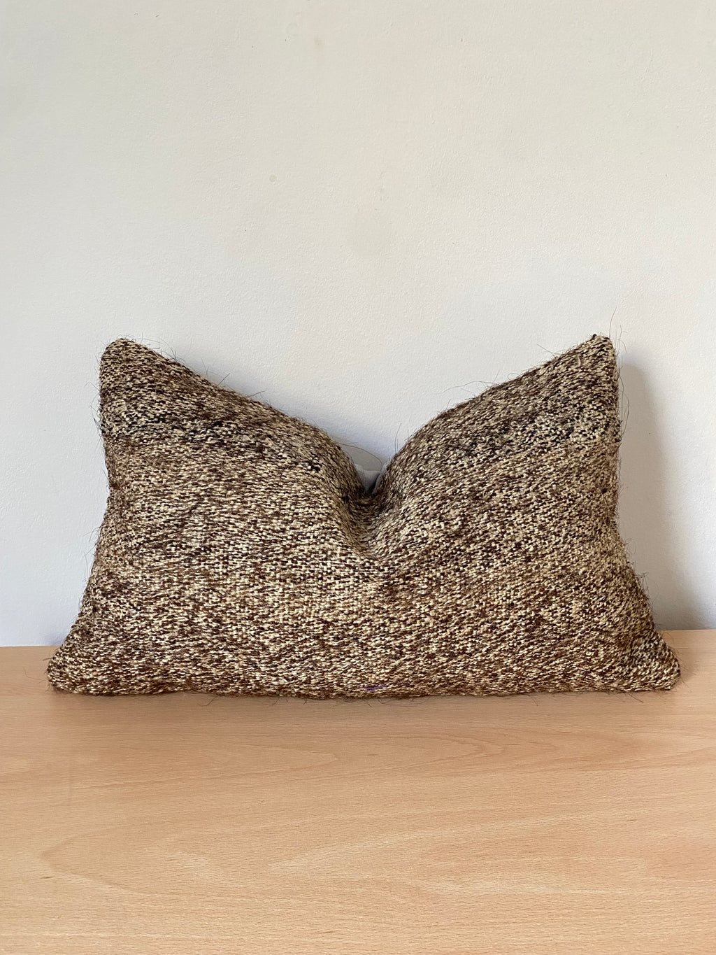 A rectangular Anatolian Turkish Kilim pillow cover in a heathered brown and beige textured fabric, resting on a light wood surface against a plain off-white wall