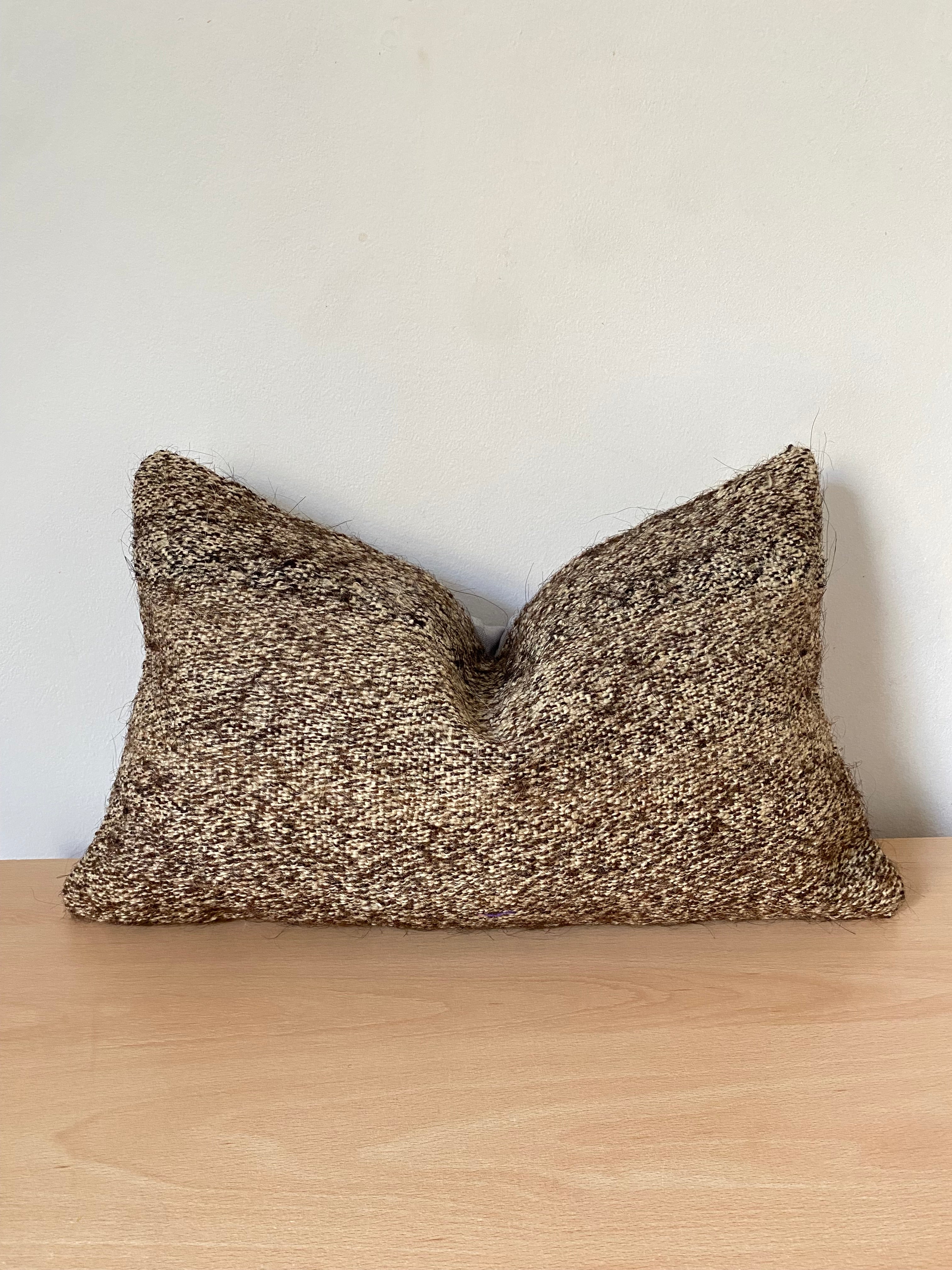 A rectangular Anatolian Turkish Kilim pillow cover in a heathered brown and beige textured fabric, resting on a light wood surface against a plain off-white wall