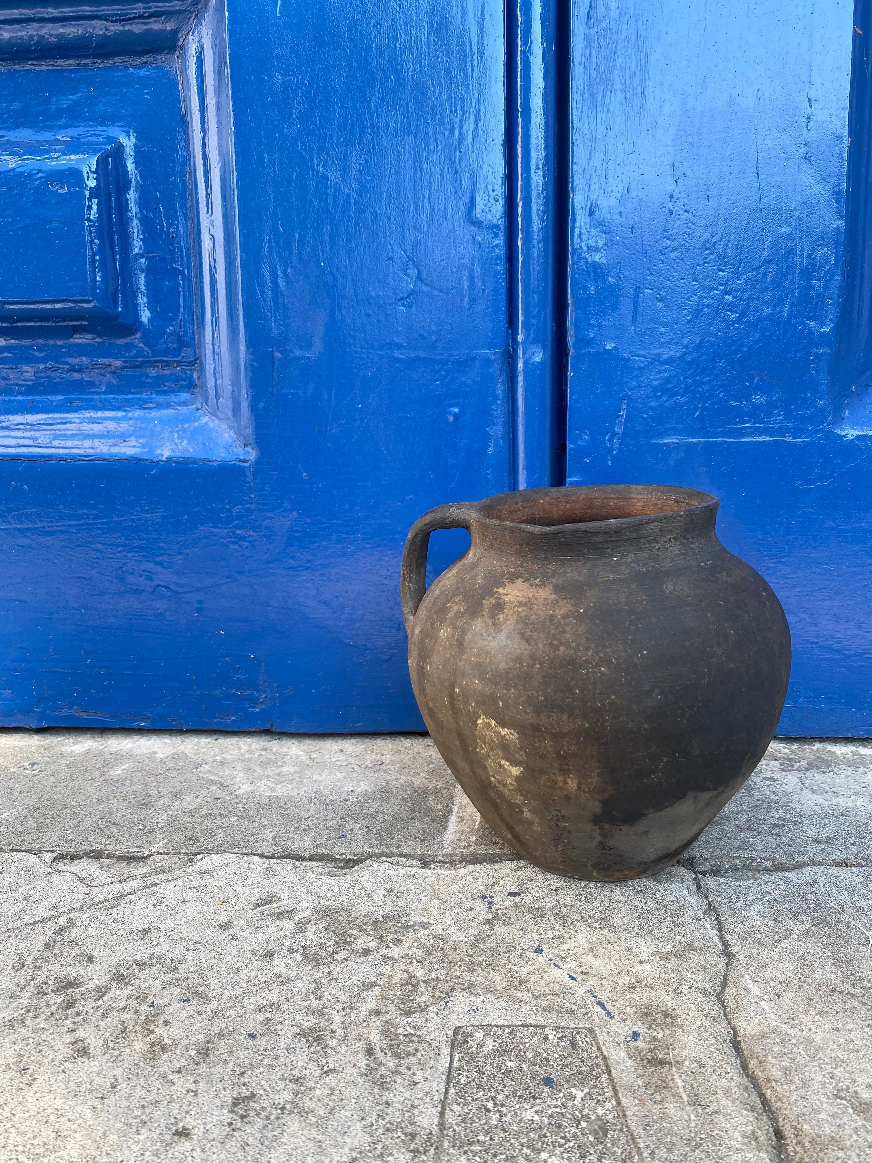 Black Antique Terracotta Clay Pot leaning against a blue door on a stone pavement