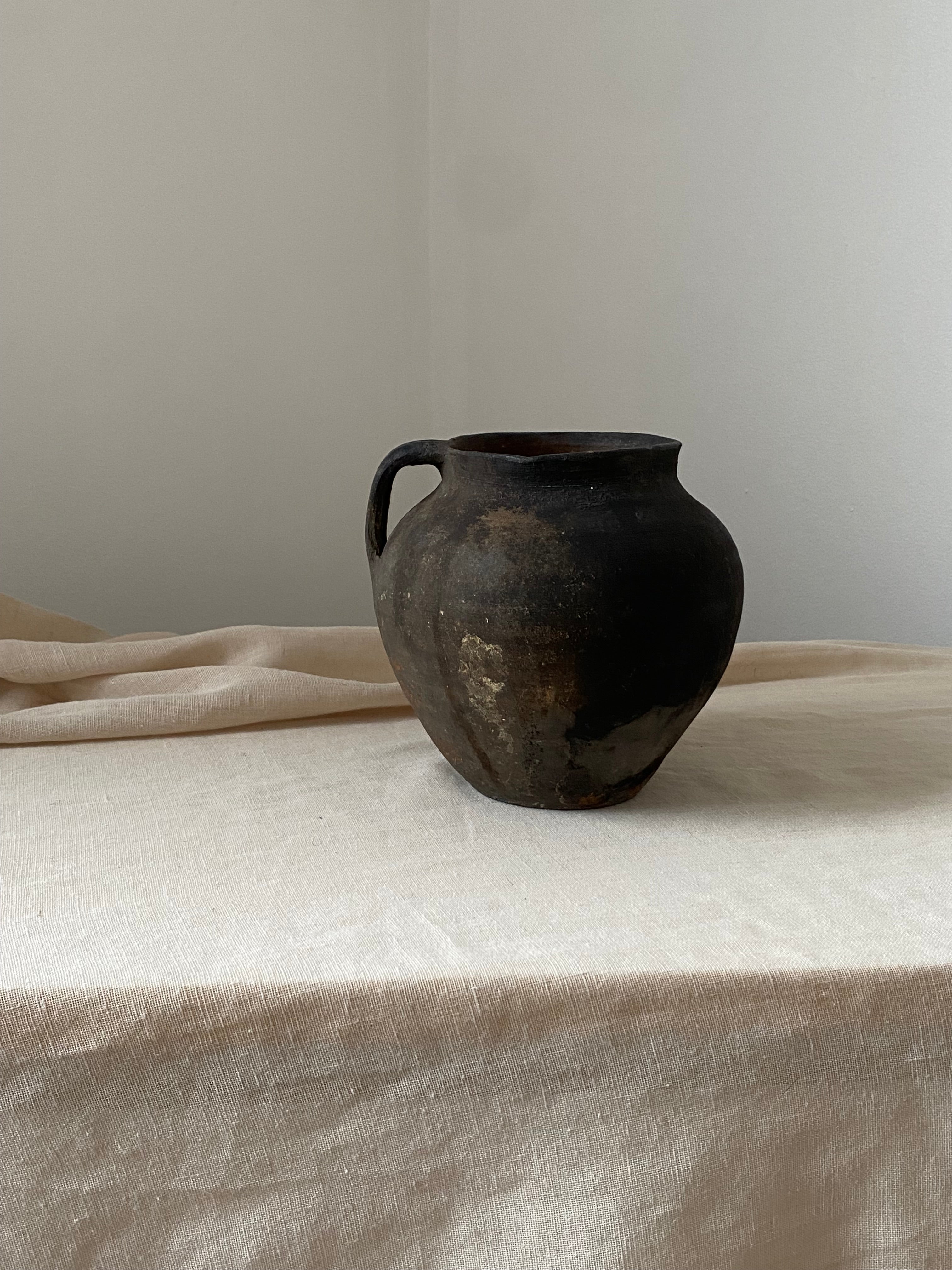 Black antique clay pot leaning against a light-colored curtain on a neutral background