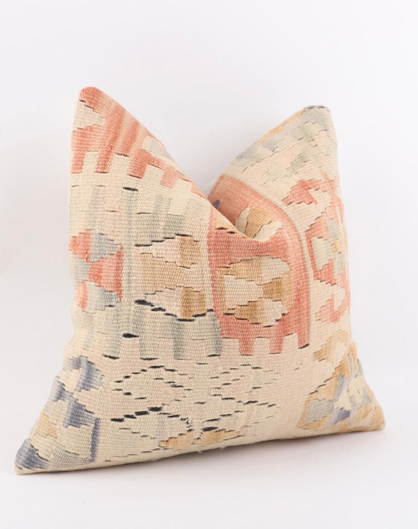 A square decorative Turkish Kilim pillow coverfeaturing a woven kilim pattern in muted tones of terracotta, beige, light blue, and grey on a cream background, photographed against a clean white studio backdrop.